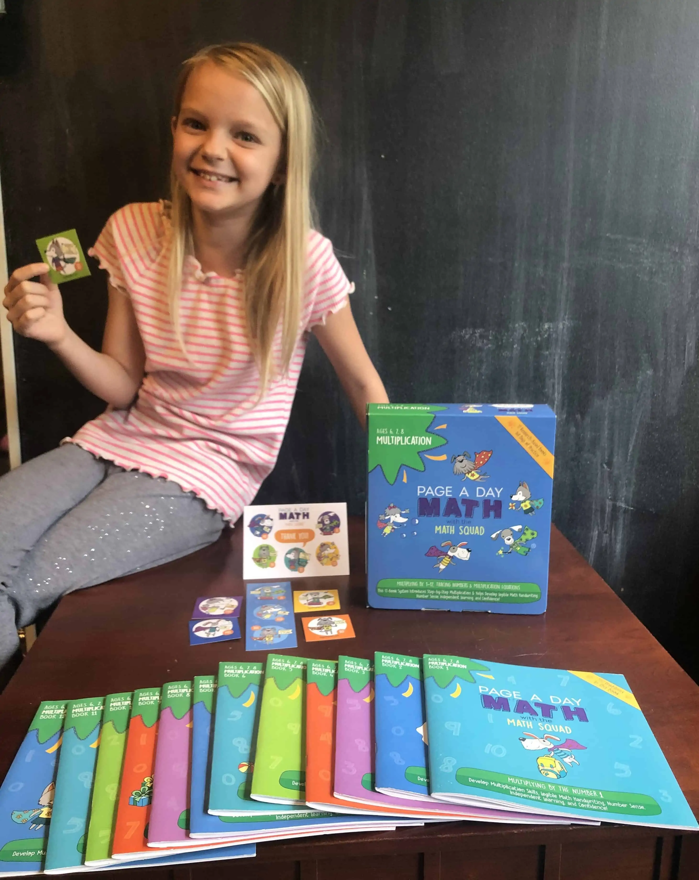 an image of a child holding a card and sitting on a table with a bunch of One Page a Day Math books shown in front of her as an example
