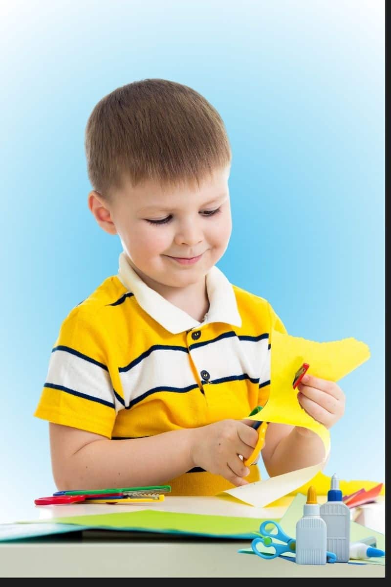 Cut and paste printables image shows a boy cutting yellow paper with other papers, scissors and glue on the table in front of him.