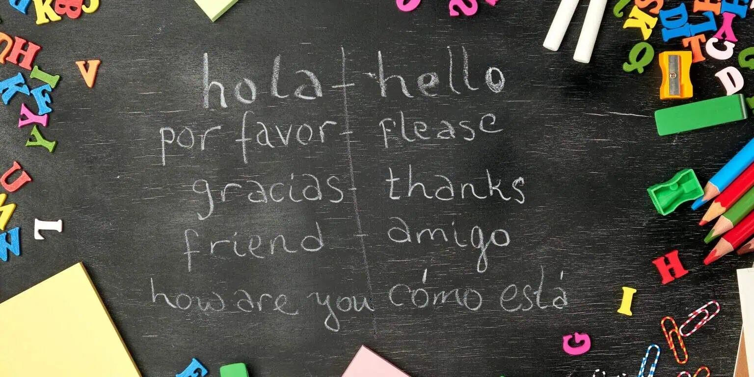Chalkboard with magnet letters, colored pencils, and paper clips. Spanish and English words like hola-hello, por favor-please, gracias-thank you, and amigo-friend are written on it, all great words that should be in a preschool Spanish curriculum.